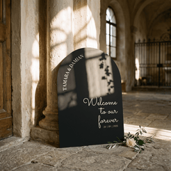 A black arched wedding welcome sign with white script text reading 'Welcome to our forever 20 | 09 | 2025' and 'TAMARA & DAMIAN' along the top edge, leaning against a stone column in a sunlit historic interior with a small white rose arrangement at the base.