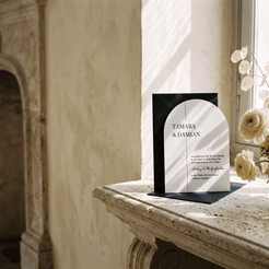 A wedding invitation card and matching envelope displayed on a stone mantelpiece in a sunlit room. The invitation features a black arch over a white background with elegant serif text reading 'TAMARA & DAMIAN' and smaller script text below.