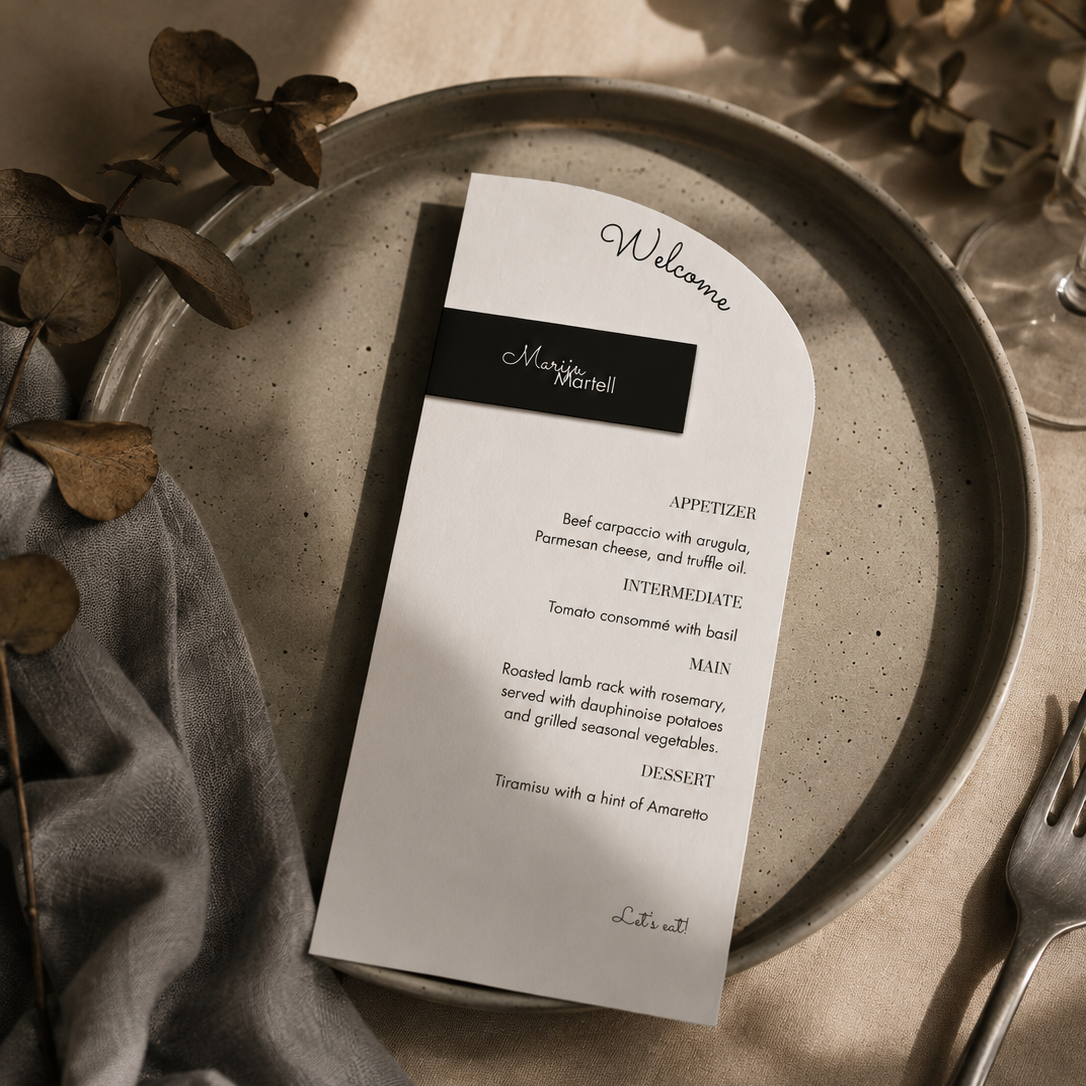 A printed wedding menu with a black name banner, styled on a speckled gray ceramic plate with a gray napkin, silver fork, and dried botanicals.
