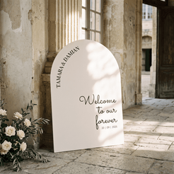 A large arched white wedding welcome sign leaning against a stone wall in an outdoor courtyard. The sign reads 'TAMARA & DAMIAN Welcome to our forever 20 | 09 | 2025' and is styled with white flowers at the base.