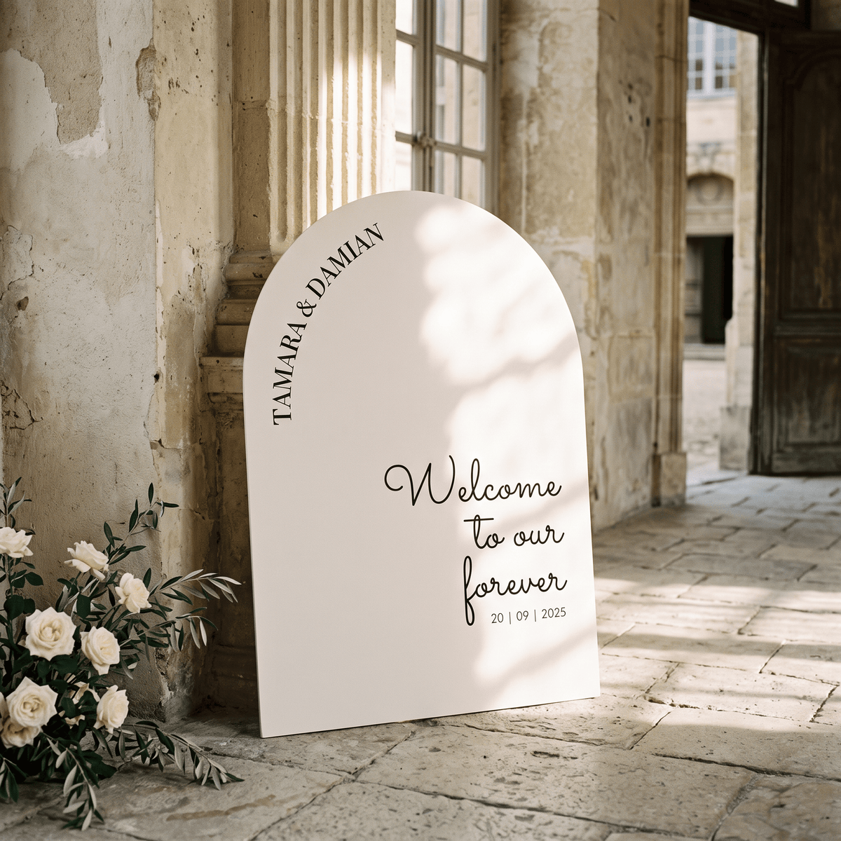 A large arched white wedding welcome sign leaning against a stone wall in an outdoor courtyard. The sign reads 'TAMARA & DAMIAN Welcome to our forever 20 | 09 | 2025' and is styled with white flowers at the base.