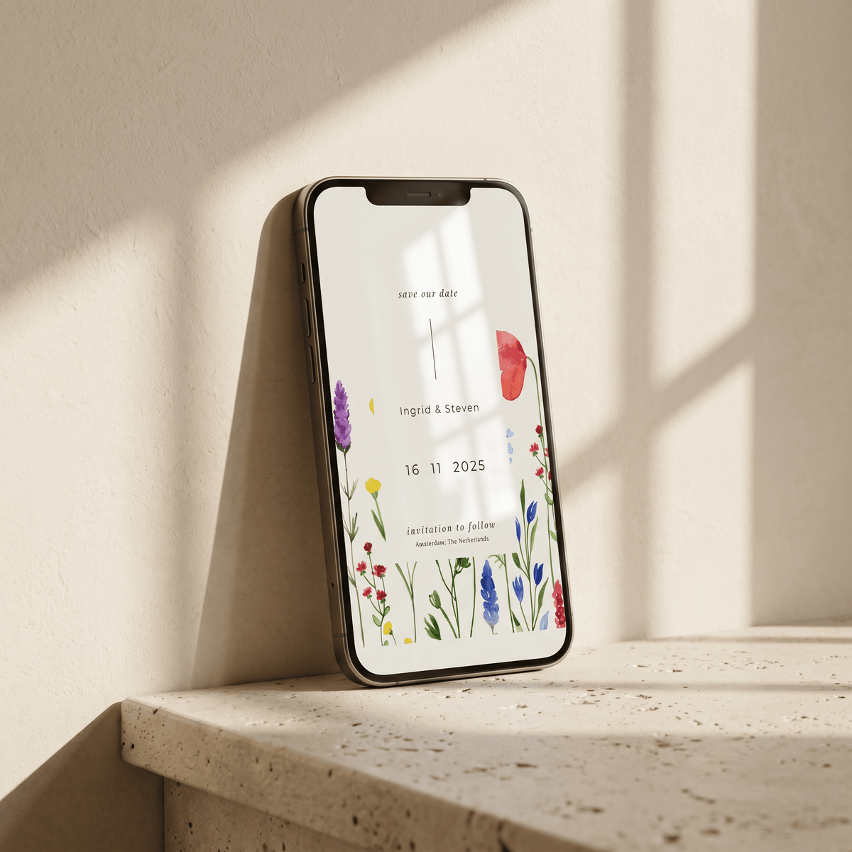 A smartphone leans against a sunlit beige wall on a stone ledge, displaying a floral 'save the date' card design.