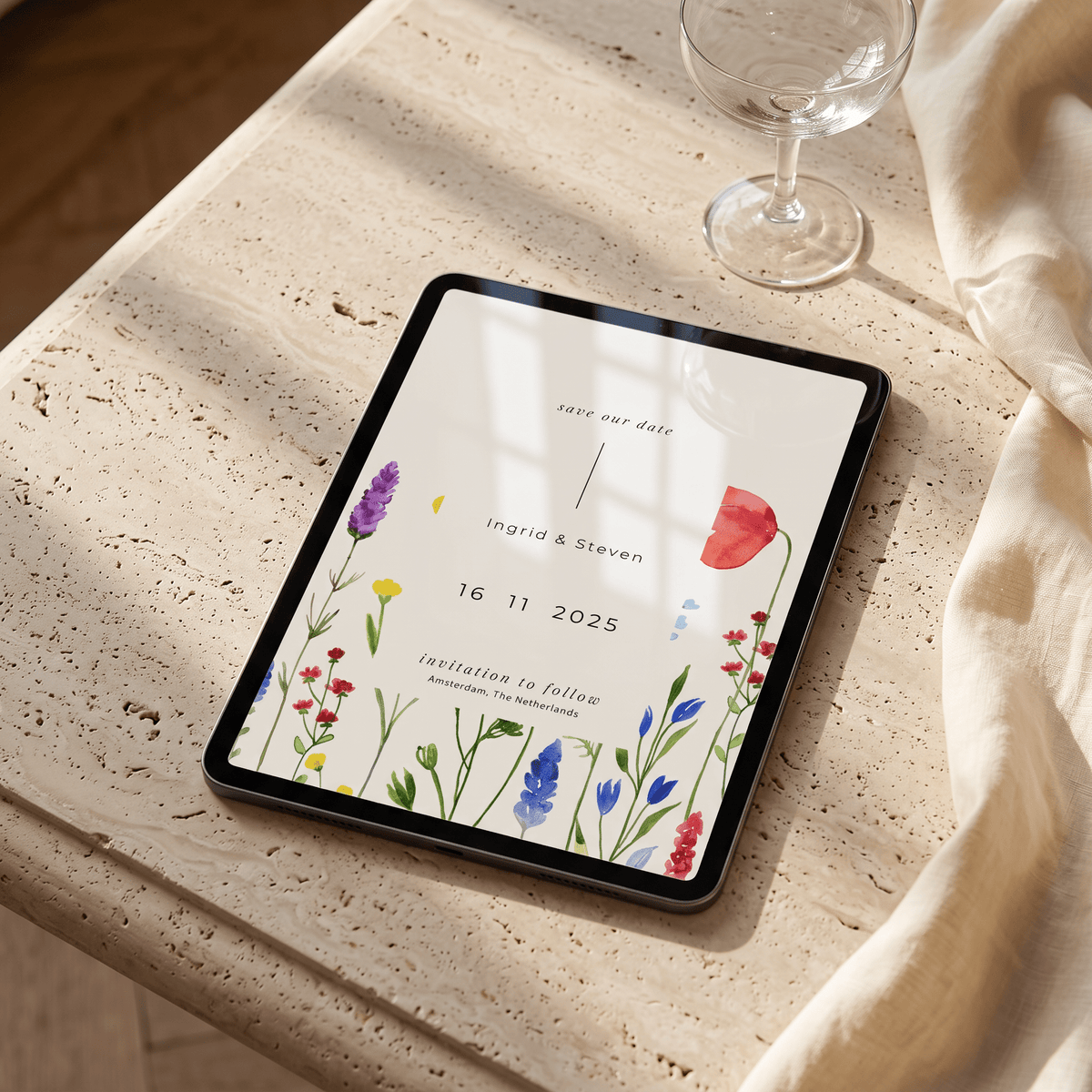 A tablet lying on a light stone tabletop, displaying a floral wedding invitation design. A wine glass and draped cream fabric are visible in the background, with warm sunlight casting shadows across the scene.