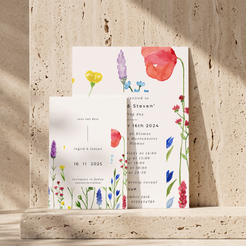 A coordinated set of floral wedding stationery cards arranged on a stone surface against a textured stone wall. The set includes a main invitation card, a save-the-date card, and a third card partially visible behind, all printed with delicate watercolor flowers in red, purple, blue, yellow, and pink on white backgrounds.