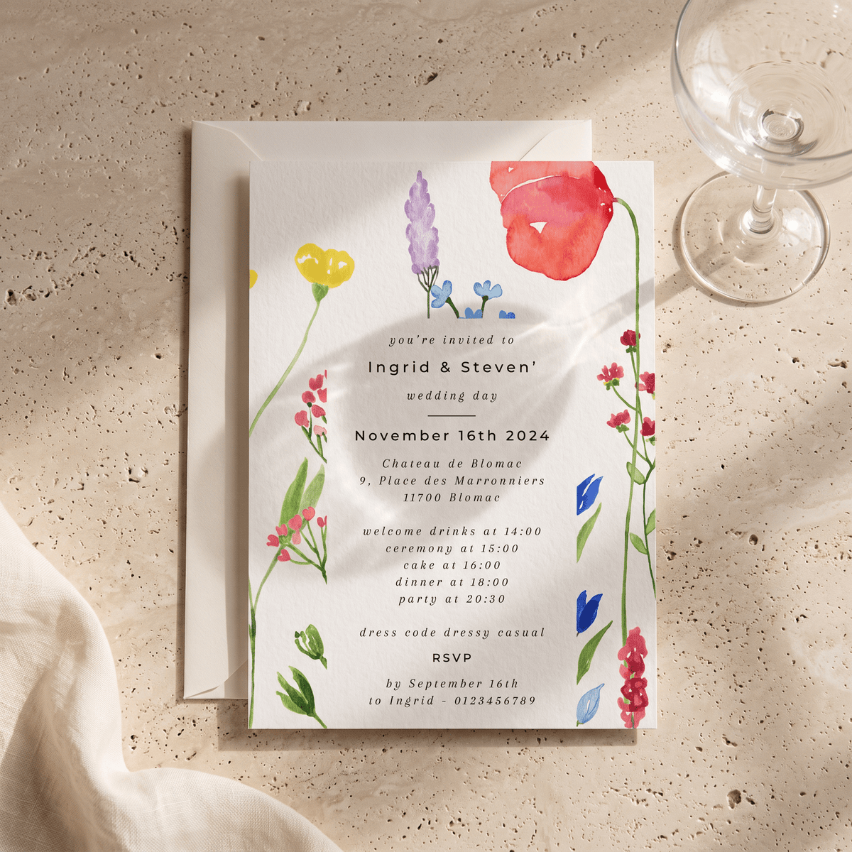 A square wedding invitation card with a white background and colorful watercolor floral illustrations, placed on top of a white envelope on a textured beige surface with a glass and fabric napkin nearby.