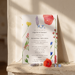 A floral wedding invitation card with elegant black typography, styled on a rustic stone ledge with soft natural shadows. The card features delicate watercolor illustrations of red, pink, purple, and yellow wildflowers with green stems on a white background.