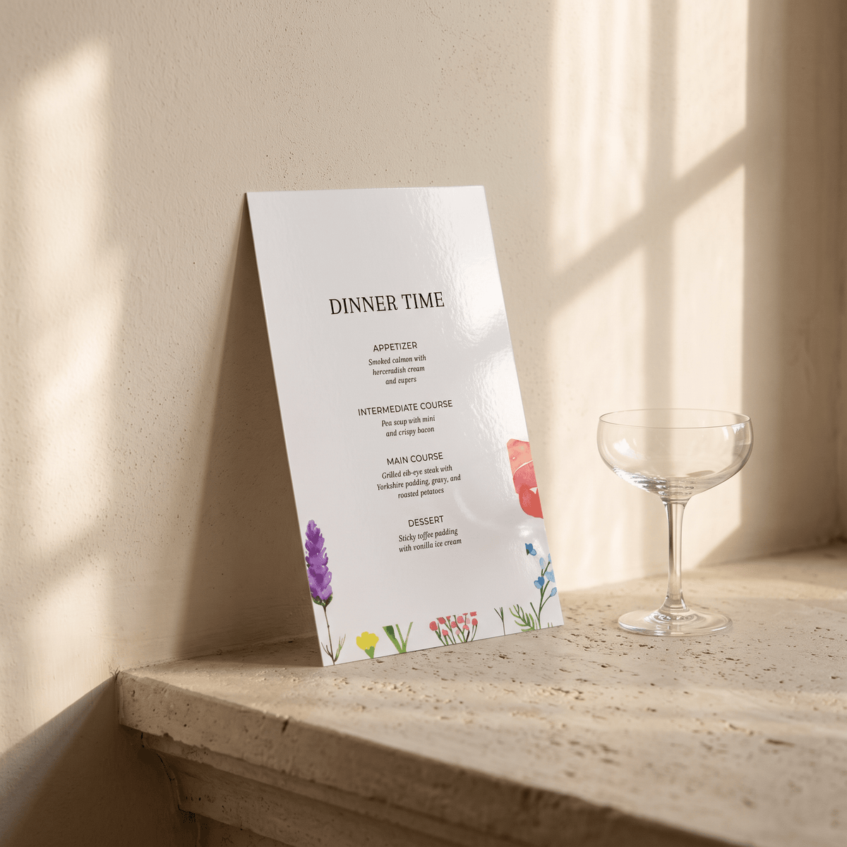 A vertical printed menu card titled "DINNER TIME" leans against a sunlit beige wall on a stone ledge, with a clear coupe glass beside it. The card is mostly white with small floral illustrations in purple, yellow, pink, and blue.