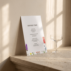 A vertical printed menu card titled "DINNER TIME" leans against a sunlit beige wall on a stone ledge, with a clear coupe glass beside it. The card is mostly white with small floral illustrations in purple, yellow, pink, and blue.
