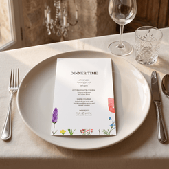 A printed dinner party menu card with floral illustrations sits on a white plate in a beautifully set dining table. The table is styled with a silver fork, silver knife, a clear glass, a wine glass, and a cream-colored tablecloth.
