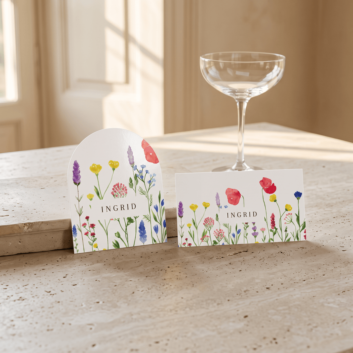 Two floral place cards with the name INGRID printed on them, styled on a light stone tabletop beside an empty coupe glass in a softly lit interior setting.