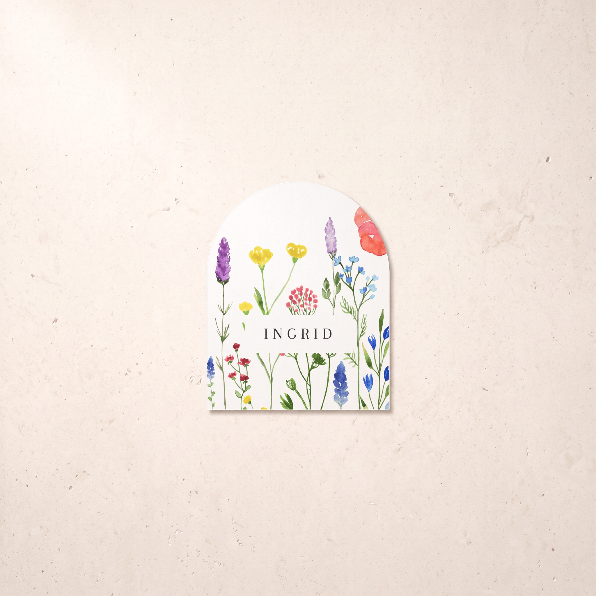 An arched white paper tag featuring a colorful watercolor illustration of wildflowers and the brand name 'INGRID' in the center, displayed on a textured beige background.