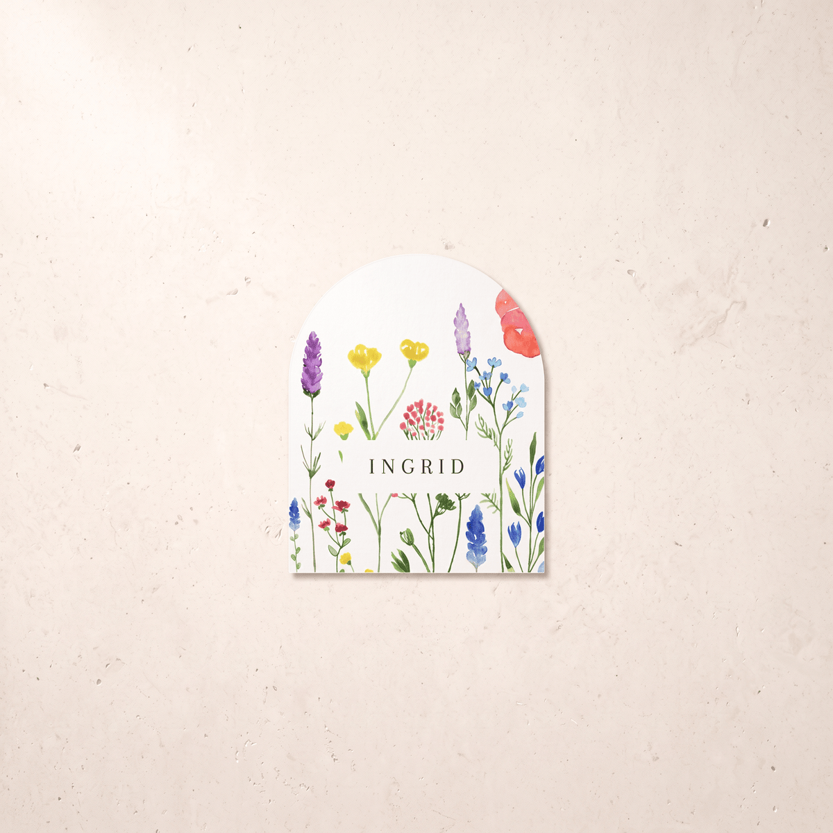 An arched white paper tag featuring a colorful watercolor illustration of wildflowers and the brand name 'INGRID' in the center, displayed on a textured beige background.