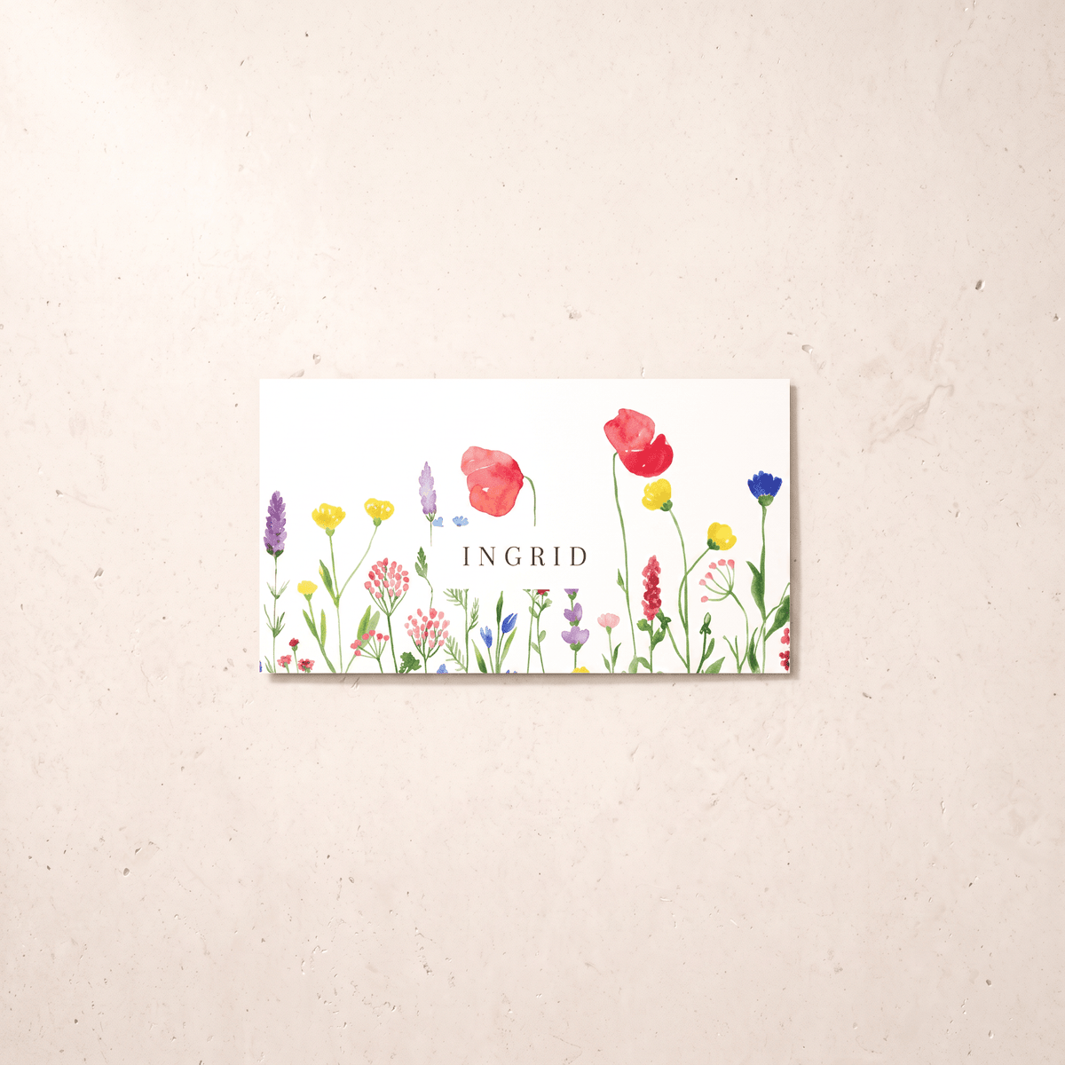 A rectangular white card featuring a colorful watercolor wildflower illustration and the name 'INGRID' in the center, displayed against a light beige textured background.