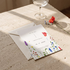 A floral wedding save-the-date card and matching envelope on a textured stone surface, with a glass and red flower nearby in soft sunlight.