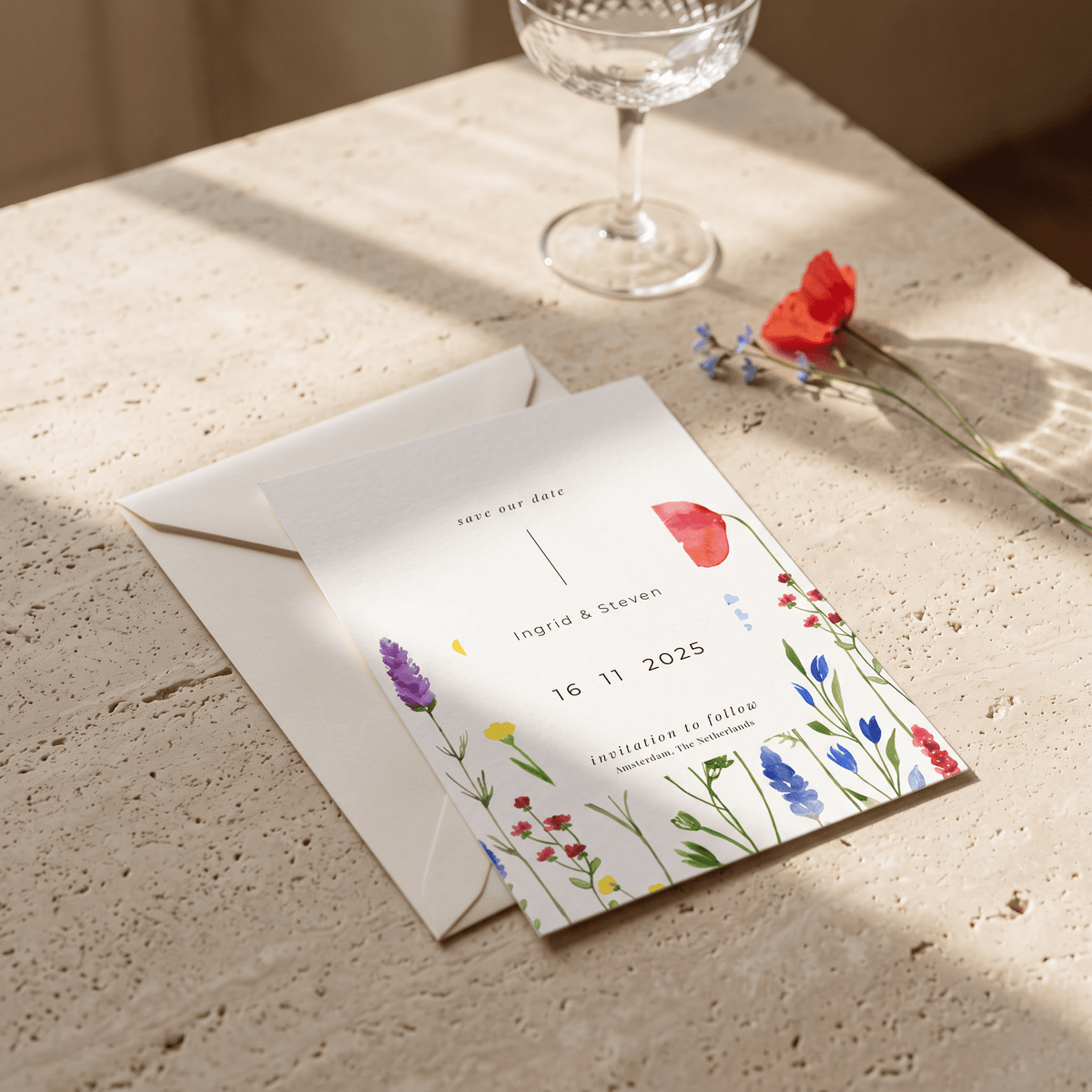 A floral wedding save-the-date card and matching envelope on a textured stone surface, with a glass and red flower nearby in soft sunlight.