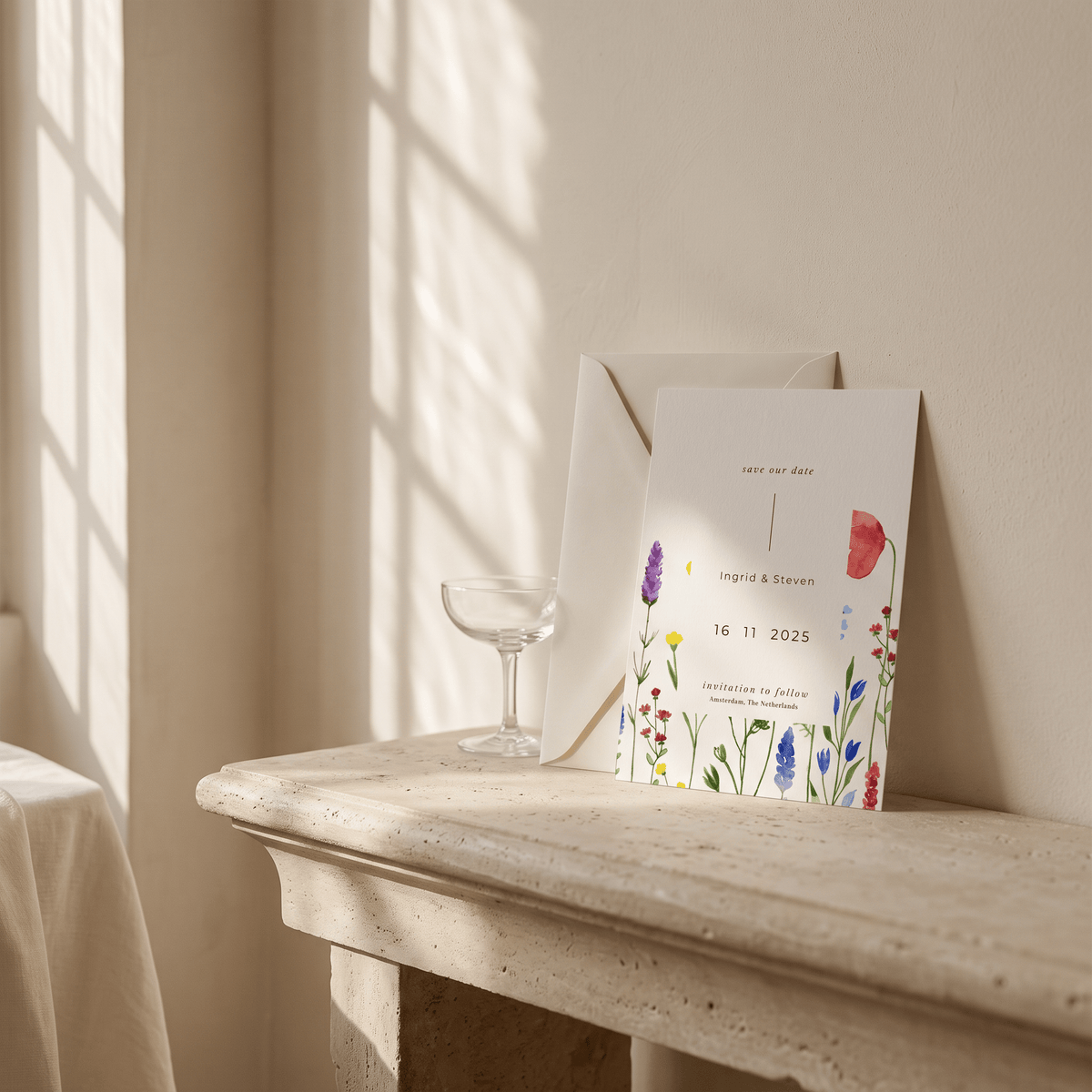 A wedding invitation card with colorful watercolor flowers and the names 'Cordelia & Steven' leans against a cream wall on a stone mantelpiece, next to a glass and an envelope in a softly lit room.
