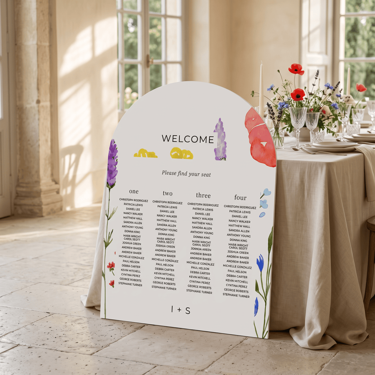 A large white wedding seating chart sign with colorful floral illustrations and printed guest names, displayed on a stone floor beside a decorated event table with flowers and glassware in a sunlit room.