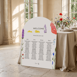 A large white wedding seating chart sign with colorful floral illustrations and printed guest names, displayed on a stone floor beside a decorated event table with flowers and glassware in a sunlit room.