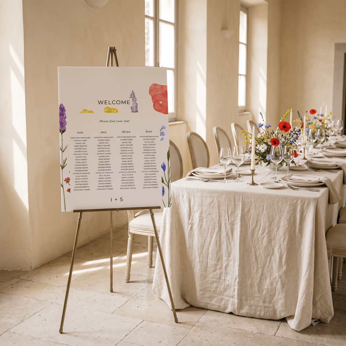 A vertical white welcome sign with colorful floral illustrations and a seated guests table layout, displayed on a gold easel in a sunlit dining room next to a linen-covered table set with flowers and glassware.