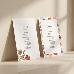 Two elegant wedding program or menu cards with floral illustrations, standing upright on a beige stone surface against a matching background.