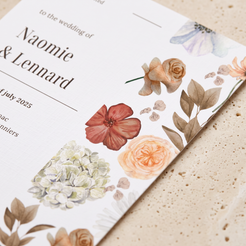 A close-up of a printed wedding invitation card with elegant black serif text and watercolor floral illustrations in muted peach, coral, cream, and taupe tones, placed on a light beige textured surface.