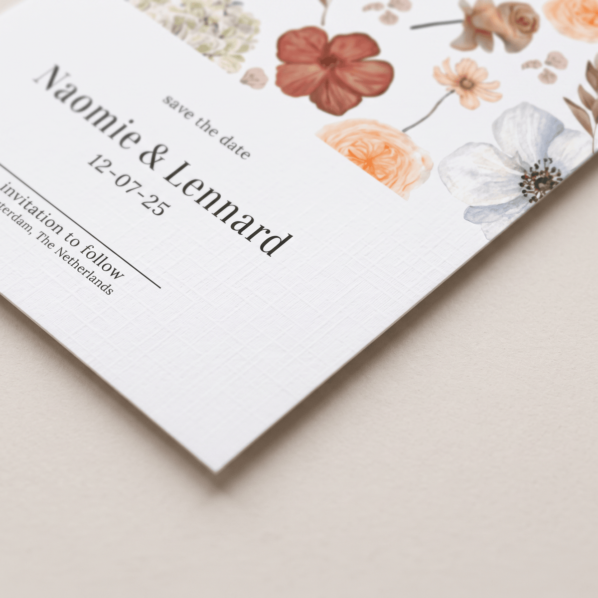 A close-up of a white wedding save-the-date card with elegant black text and delicate watercolor floral illustrations in peach, pink, and soft blue, resting on a neutral beige surface.
