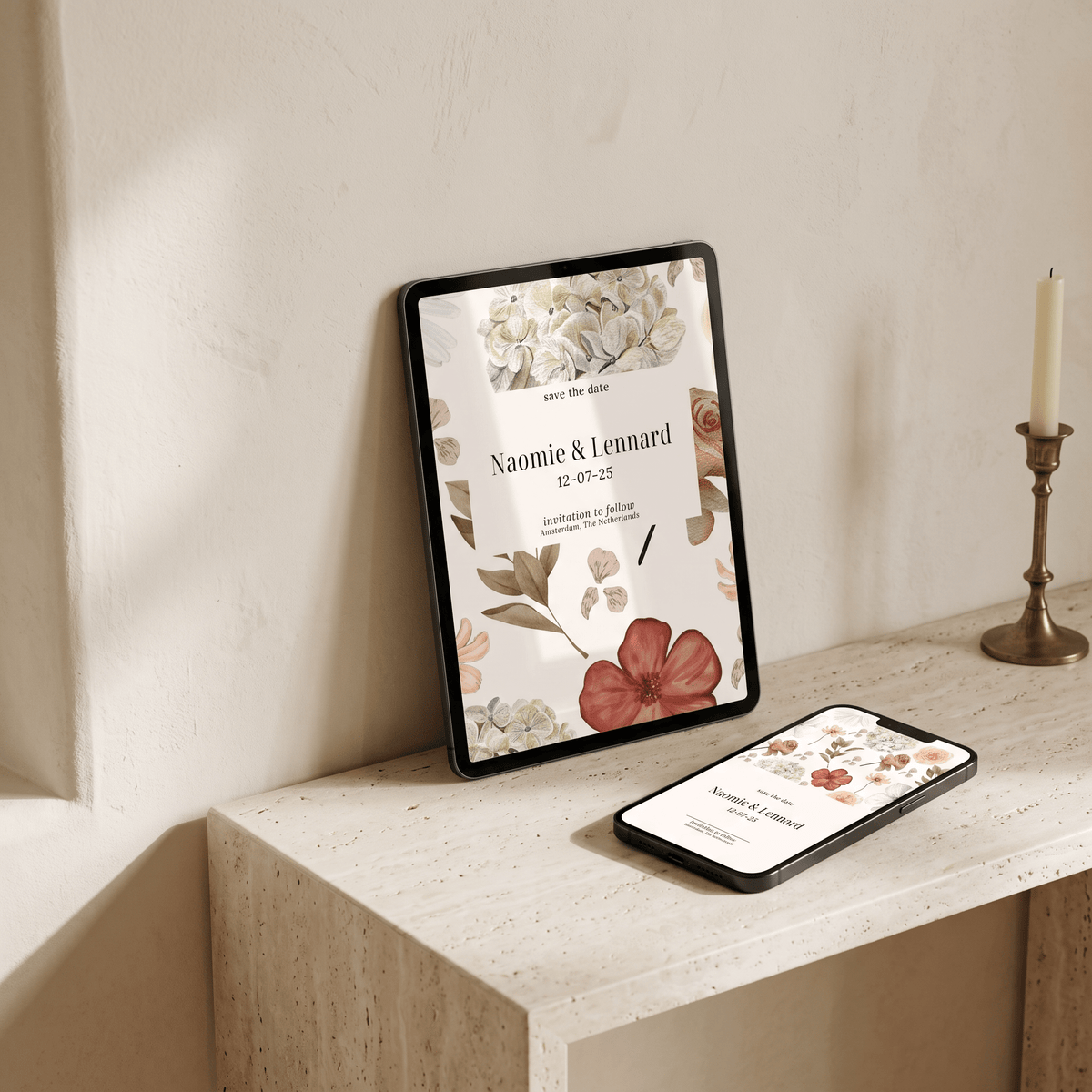 A tablet and smartphone displaying matching floral invitation designs rest on a light stone surface against a warm beige wall, with a candle in a brass holder nearby.