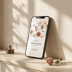 A smartphone leaning against a textured cream wall on a stone ledge, displaying a floral save-the-date invitation design. The scene is styled with warm natural shadows, a small brass object, and a dried rose in the foreground.