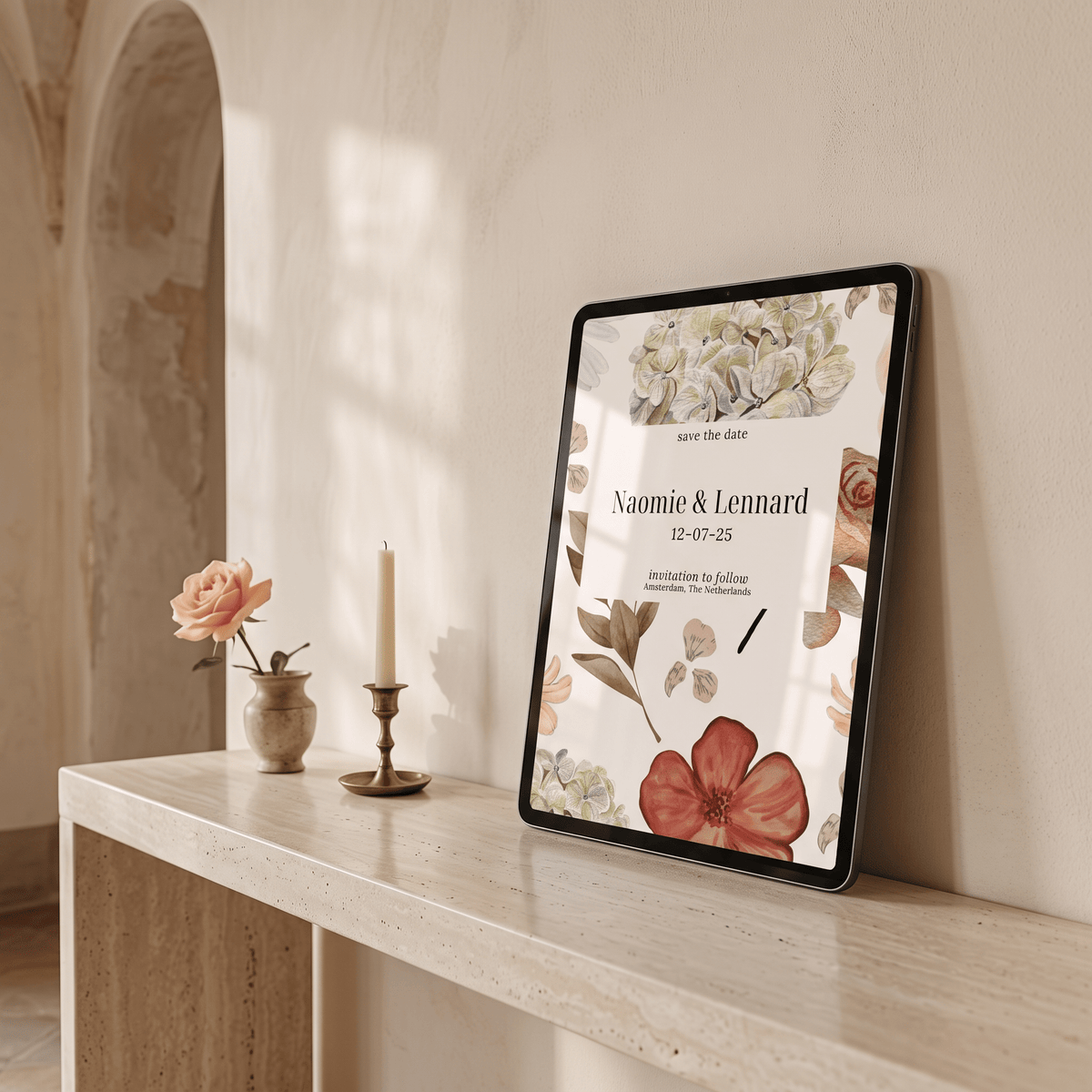 A framed floral 'save the date' invitation card leans against a cream wall on a light stone console table, styled with a small vase of peach flower and a candle in a softly lit interior.