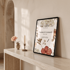 A framed floral 'save the date' invitation card leans against a cream wall on a light stone console table, styled with a small vase of peach flower and a candle in a softly lit interior.