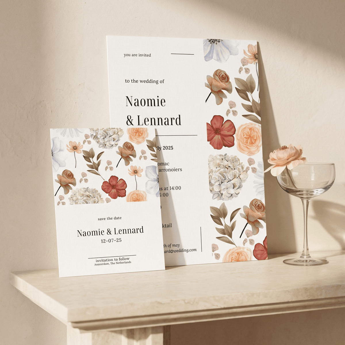Floral wedding invitation stationery set displayed on a light stone table against a warm beige wall, with a wine glass holding a flower beside it. The set includes a main invitation card, a save-the-date card, and matching floral border designs.