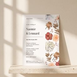 A floral wedding invitation card displayed on a stone ledge against a warm beige wall with soft window shadows. The card features elegant black typography on a white background with watercolor-style flowers in peach, coral, cream, and muted blue along the right side.