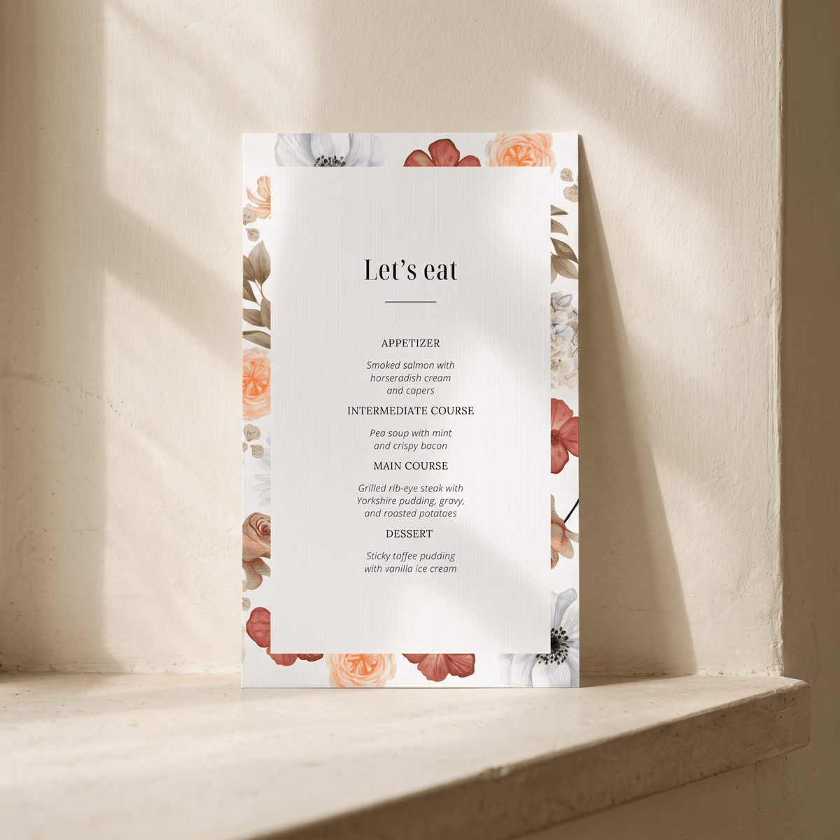 A vertical wedding program or menu card with a white center and floral border, standing on a stone ledge against a textured beige wall with soft sunlight and shadows.