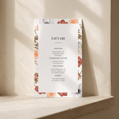 A vertical wedding program or menu card with a white center and floral border, standing on a stone ledge against a textured beige wall with soft sunlight and shadows.