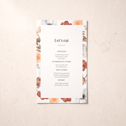 A vertical wedding menu card with a white center and a floral border in muted peach, dusty rose, and sage tones, photographed on a light beige textured surface. The printed text reads “Let’s eat” with course suggestions below.