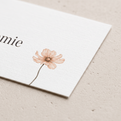 A close-up of a white textured card with part of the word 'emie' printed in black and a delicate pink flower illustration, resting on a light beige textured surface.