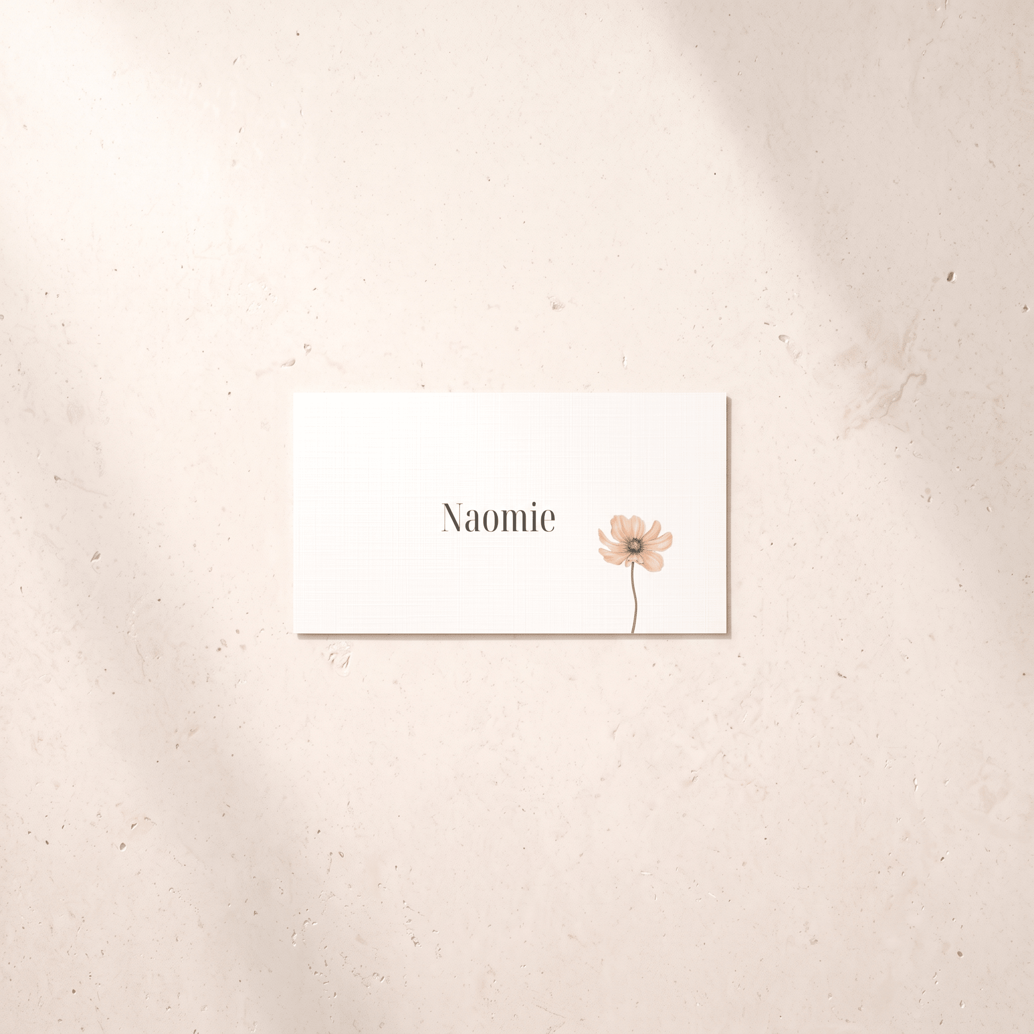 A rectangular white card with the name 'Naomie' in black serif text and a small pale pink flower illustration, placed on a light beige textured surface with soft shadows.