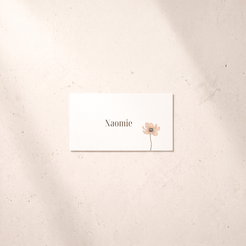 A rectangular white card with the name 'Naomie' in black serif text and a small pale pink flower illustration, placed on a light beige textured surface with soft shadows.