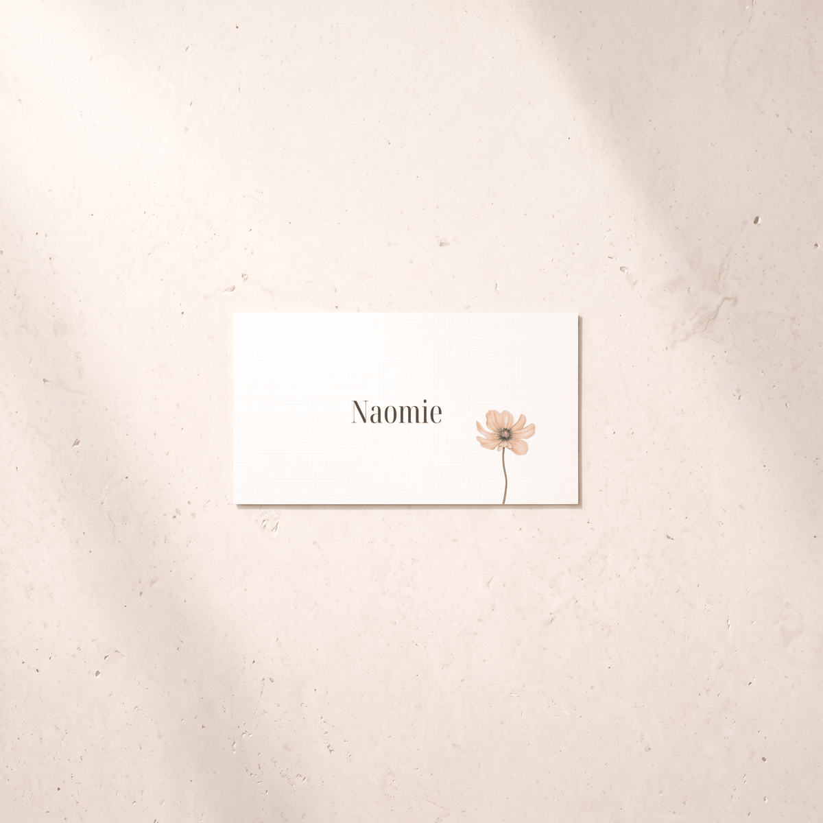 A rectangular white card with the name 'Naomie' in black serif text and a small pale pink flower illustration, placed on a light beige textured surface with soft shadows.