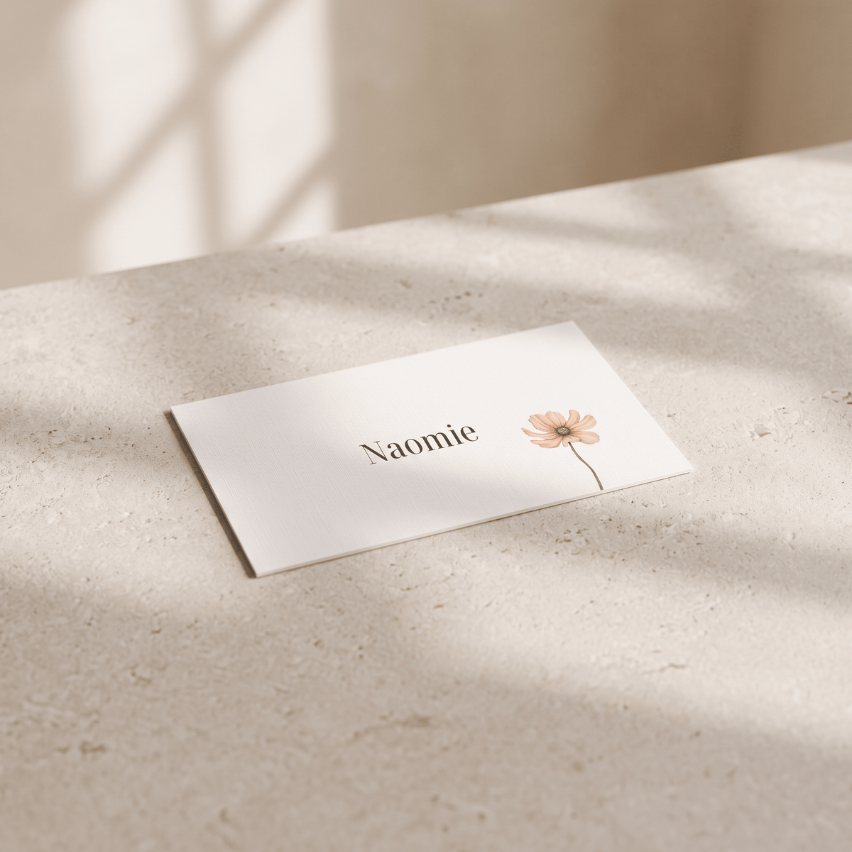 A white rectangular name card with the name 'Naomie' in black serif text and a small peach flower illustration, placed on a light beige stone surface with soft window shadows.