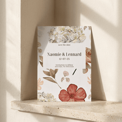 A vertical floral 'save the date' card propped against a textured cream wall on a stone ledge. The card has botanical illustrations in soft green, peach, and rust tones with a central white panel reading 'save the date', 'Naomie & Lennard', and the date '12-07-25'.