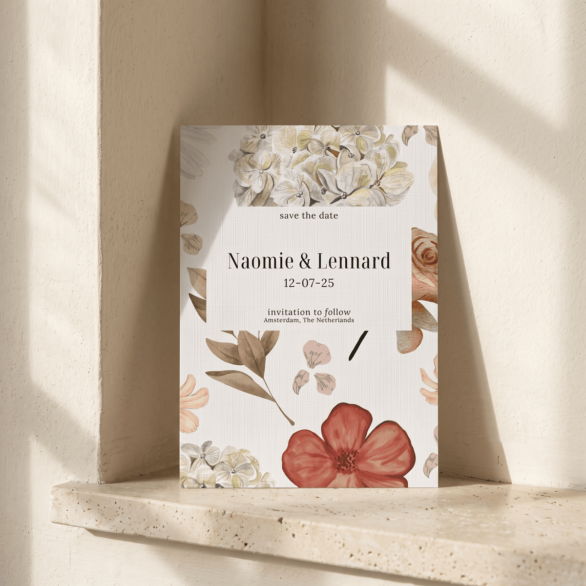 A vertical floral 'save the date' card propped against a textured cream wall on a stone ledge. The card has botanical illustrations in soft green, peach, and rust tones with a central white panel reading 'save the date', 'Naomie & Lennard', and the date '12-07-25'.