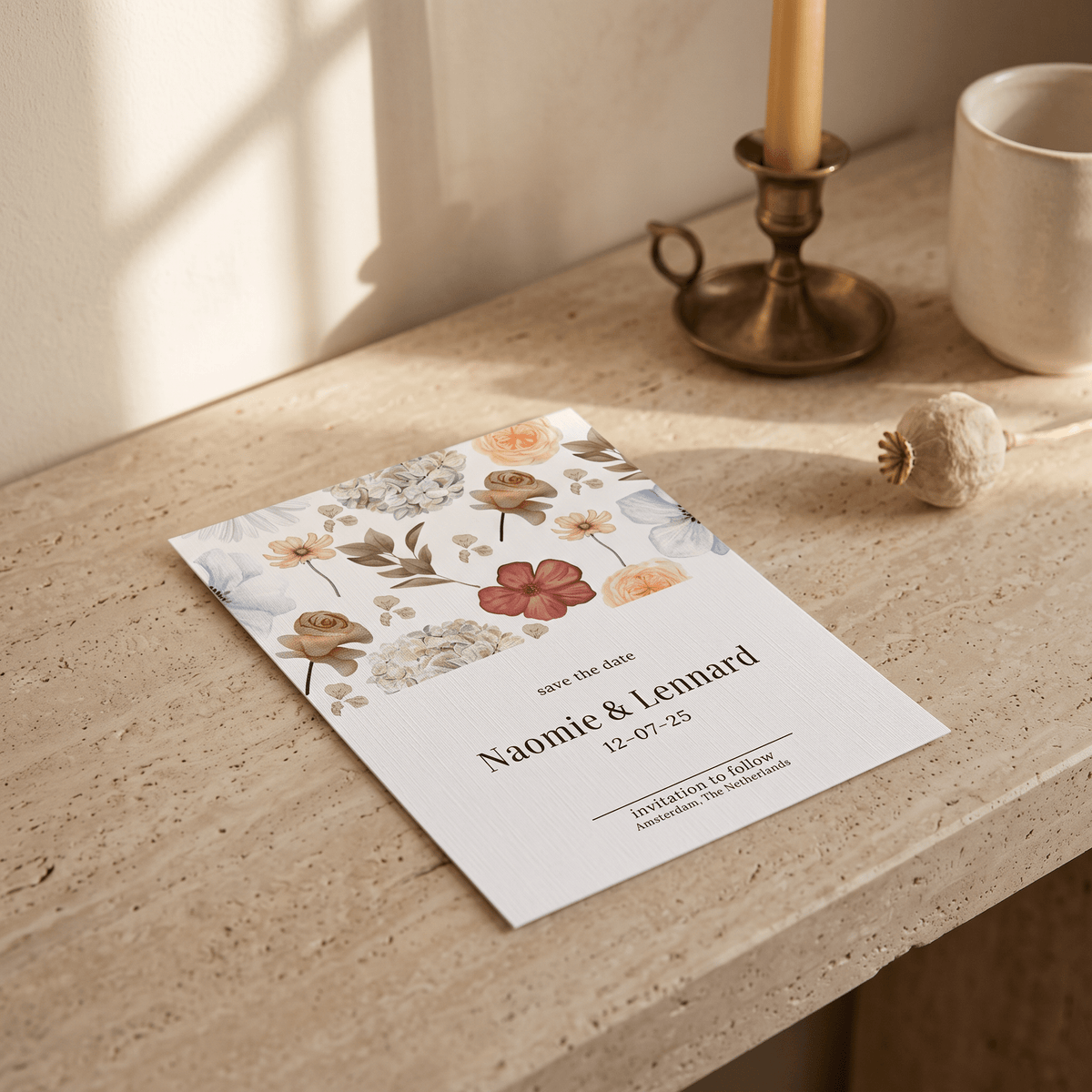 A floral 'Save the Date' card lies on a beige stone surface in a softly lit interior setting, with a brass candlestick, a ceramic mug, and a dried flower nearby. The card features pastel watercolor flowers and the names Naomi & Lennard with the date 12.
