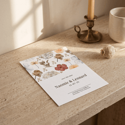 A floral 'Save the Date' card lies on a beige stone surface in a softly lit interior setting, with a brass candlestick, a ceramic mug, and a dried flower nearby. The card features pastel watercolor flowers and the names Naomi & Lennard with the date 12.