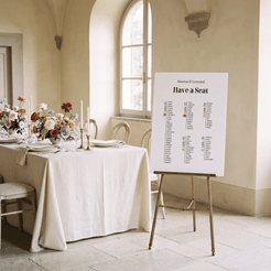 A wedding seating chart printed on a white board with black text, displayed on a gold metal easel in a bright, elegant room with beige walls and stone floors. The background shows a beautifully set dining table with white linens, floral arrangements, and glassware.