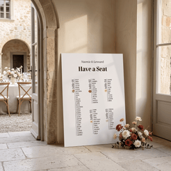 A large white wedding seating chart print leaning against a stone wall in a sunlit courtyard. The print includes the names 'Naomie & Leonard' at the top and table assignments from table 1 to table 4 in black text, with a small red rose graphic and a bouquet of flowers placed beside it.