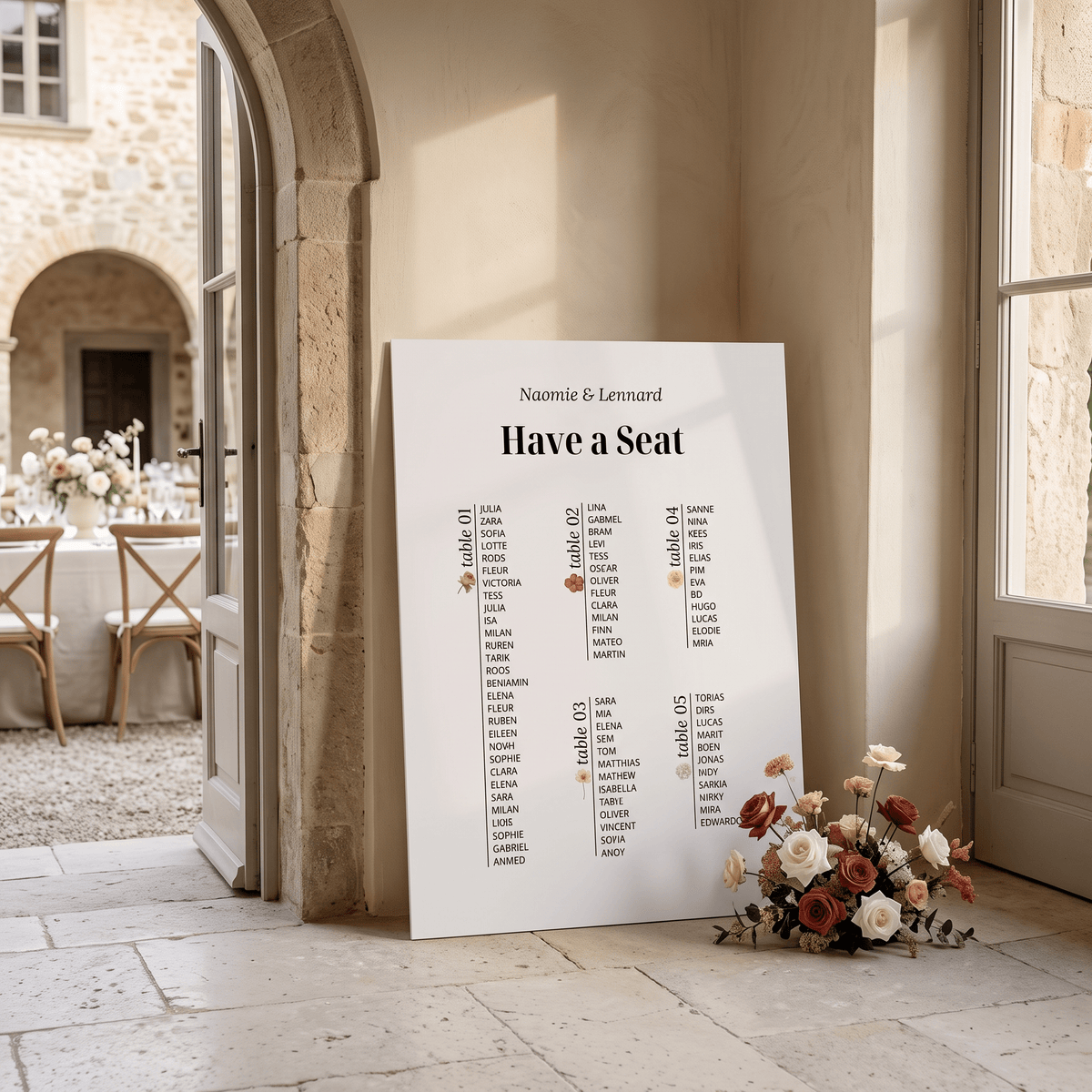 A large white wedding seating chart print leaning against a stone wall in a sunlit courtyard. The print includes the names 'Naomie & Leonard' at the top and table assignments from table 1 to table 4 in black text, with a small red rose graphic and a bouquet of flowers placed beside it.