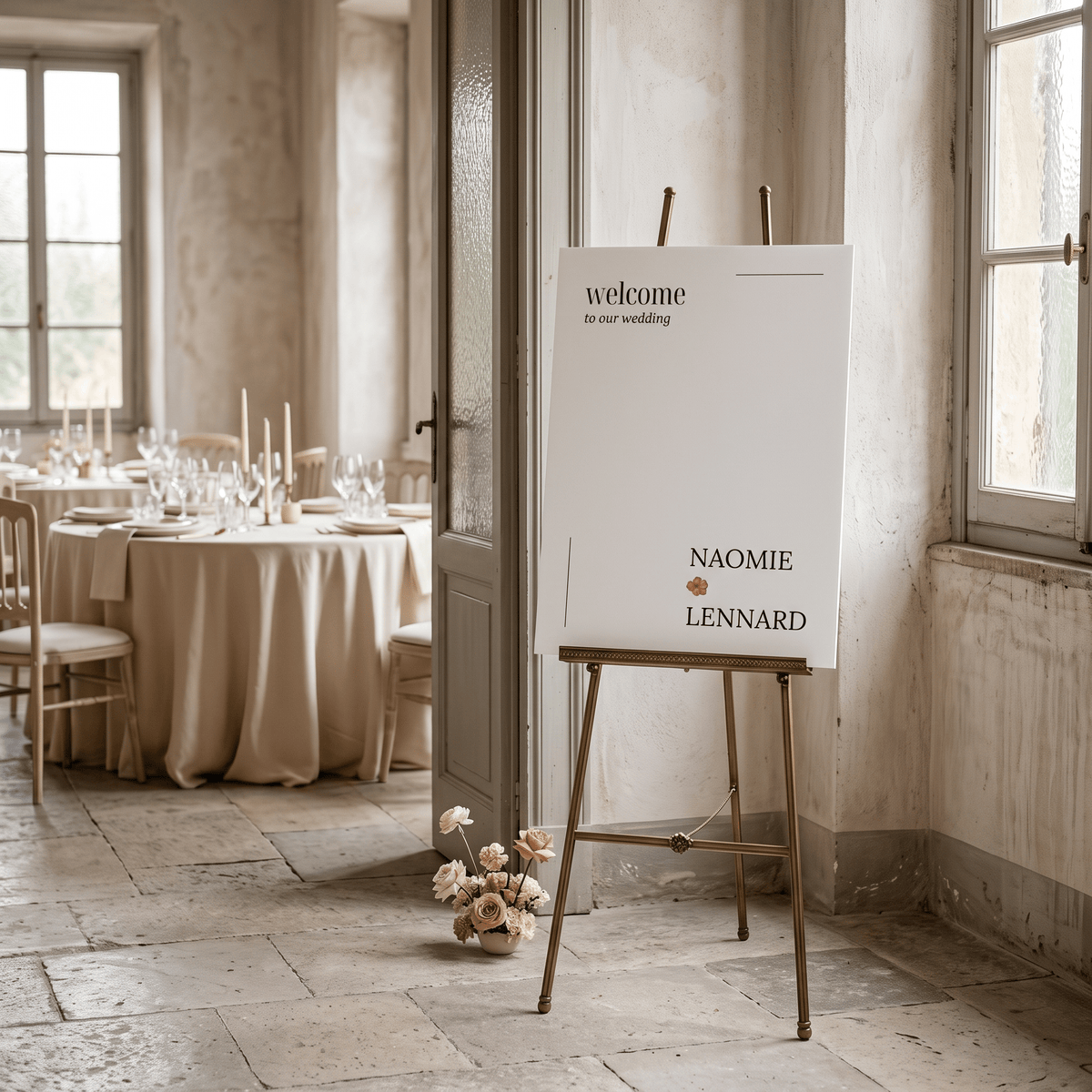 A white wedding welcome sign on a gold easel in a rustic, elegant event space with stone floors and tables set for guests.