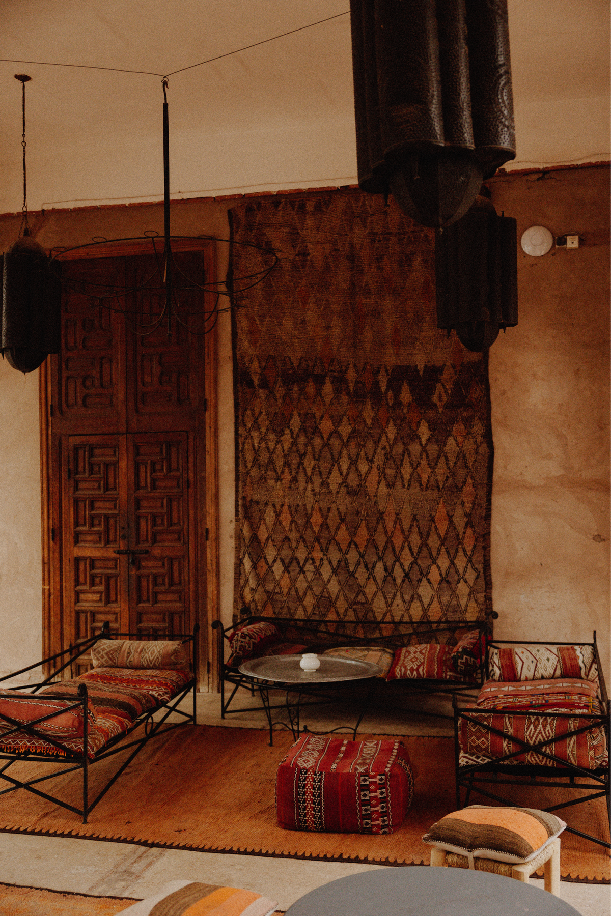 A styled Moroccan-style interior featuring a large patterned rug hanging on the wall, two black metal lounge chairs with red and beige patterned cushions, a small round coffee table, and a red patterned pouf on a wooden floor.