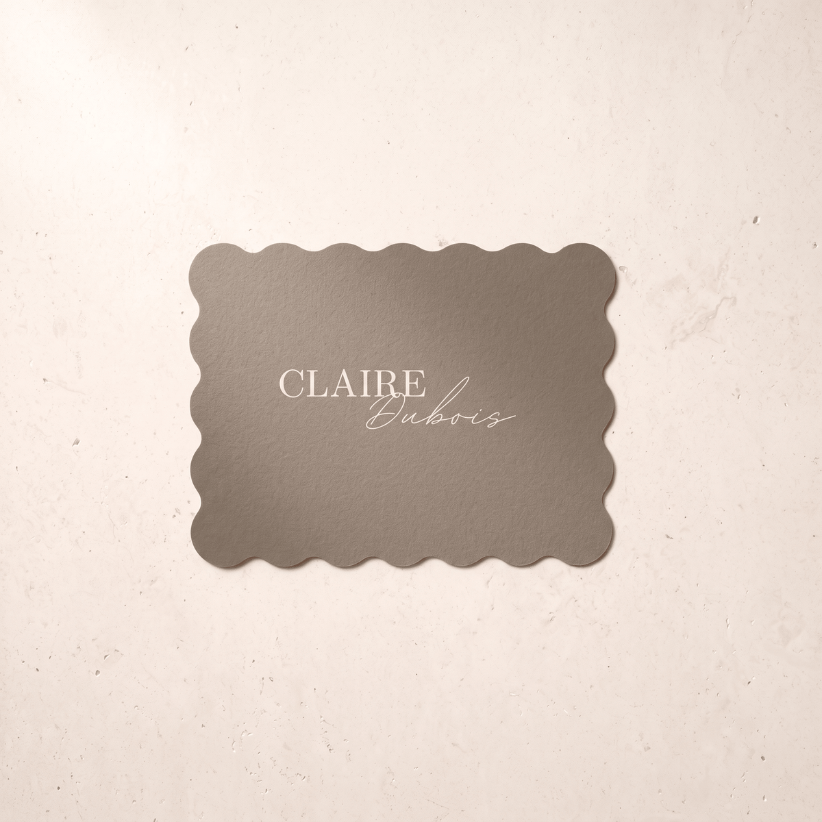 A rectangular scalloped-edge card or label in warm taupe with the text 'CLAIRE Dubois' printed in white, placed on a light beige textured surface.